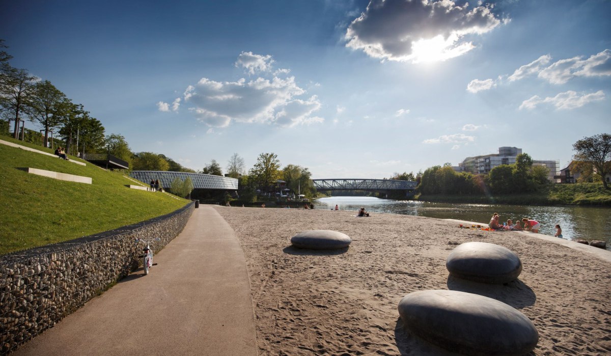 Sonniger Tag am Flussufer mit Sandstrand, großen Steinen und einer Brücke im Hintergrund. Menschen entspannen am Wasser., © Stoppel Sonniger Tag am Flussufer mit Sandstrand, großen Steinen und einer Brücke im Hintergrund. Menschen entspannen am Wasser., © Stoppel