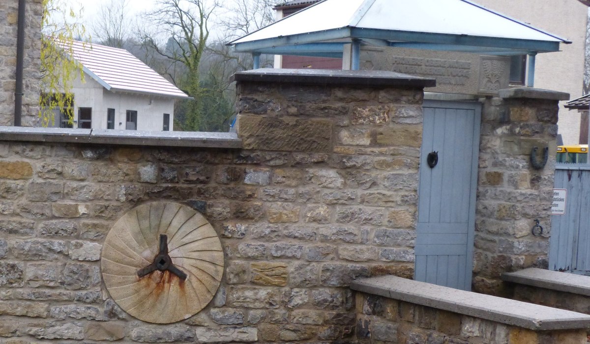 Steinmauer mit integriertem Mühlstein und blauem Holztor, überdacht von einem kleinen Dach. Im Hintergrund Bäume und Gebäude., © Land der 1000 Hügel - Kraichgau-Stromberg