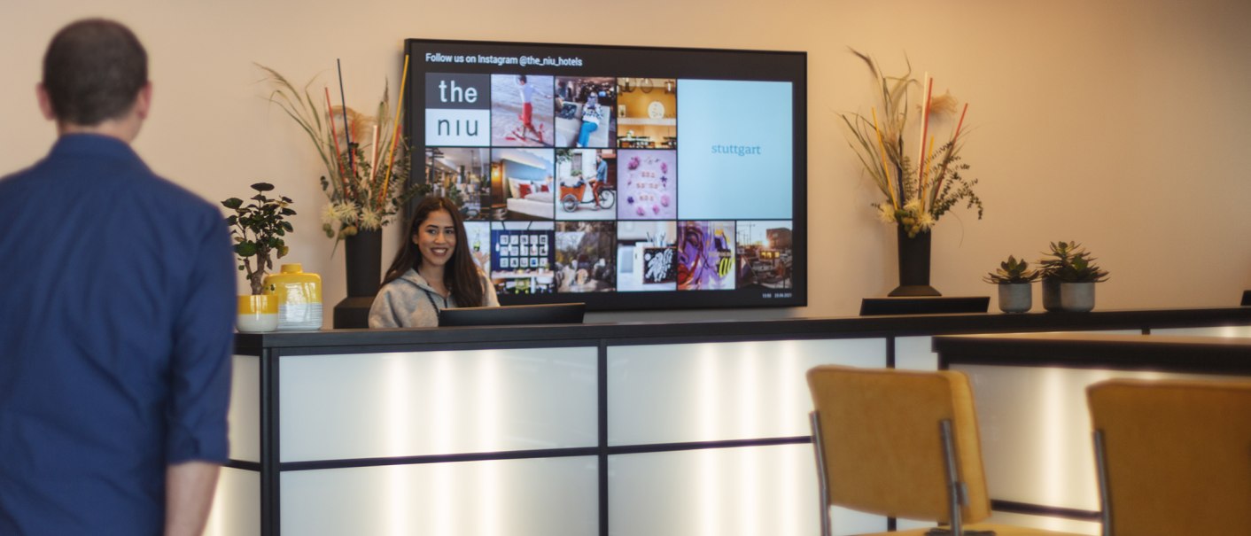 Eine Frau steht lächelnd an einer Hotelrezeption. Im Hintergrund ist ein Bildschirm mit Bildern und Texten zu sehen. Dekorationen und Pflanzen schmücken den Tresen., © the niu Form - Novum Hospitality