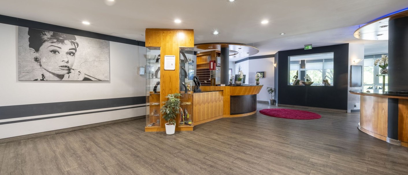 Moderne Empfangslobby mit Holzmöbeln, einem großen Schwarz-Weiß-Porträt an der Wand und einem roten Teppich auf dem Boden., © Plaza INN Stuttgart-Filderstadt Moderne Empfangslobby mit Holzmöbeln, einem großen Schwarz-Weiß-Porträt an der Wand und einem roten Teppich auf dem Boden., © Plaza INN Stuttgart-Filderstadt