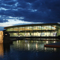 Das Oskar Frech SeeBad bei Nacht, mit einem beleuchteten Felsen im Wasser und einem modernen, verglasten Gebäude im Hintergrund., © SMG Das Oskar Frech SeeBad bei Nacht, mit einem beleuchteten Felsen im Wasser und einem modernen, verglasten Gebäude im Hintergrund., © SMG