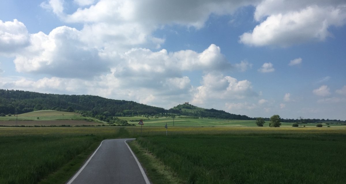 Ein Radweg schlängelt sich durch grüne Felder, im Hintergrund die Wurmlinger Kapelle auf einem Hügel unter einem bewölkten Himmel., © www.pro-cycl.de Ein Radweg schlängelt sich durch grüne Felder, im Hintergrund die Wurmlinger Kapelle auf einem Hügel unter einem bewölkten Himmel., © www.pro-cycl.de