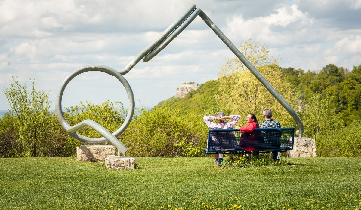 Drei Personen sitzen auf einer Bank vor einer großen Brillenskulptur in einer grünen Landschaft mit Hügeln und Wolken am Himmel., © hochgehberge Drei Personen sitzen auf einer Bank vor einer großen Brillenskulptur in einer grünen Landschaft mit Hügeln und Wolken am Himmel., © hochgehberge