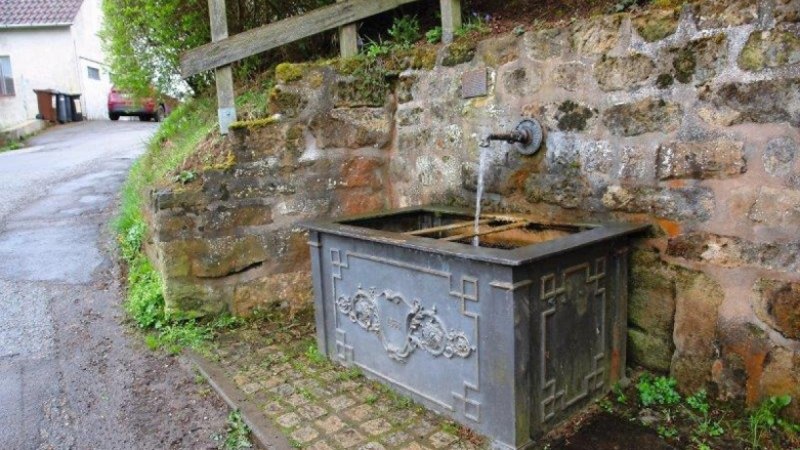 Ein historischer Brunnen mit fließendem Wasser, eingebaut in eine Steinmauer. Der Brunnen hat dekorative Verzierungen., © Bürgerausschuss WHSO