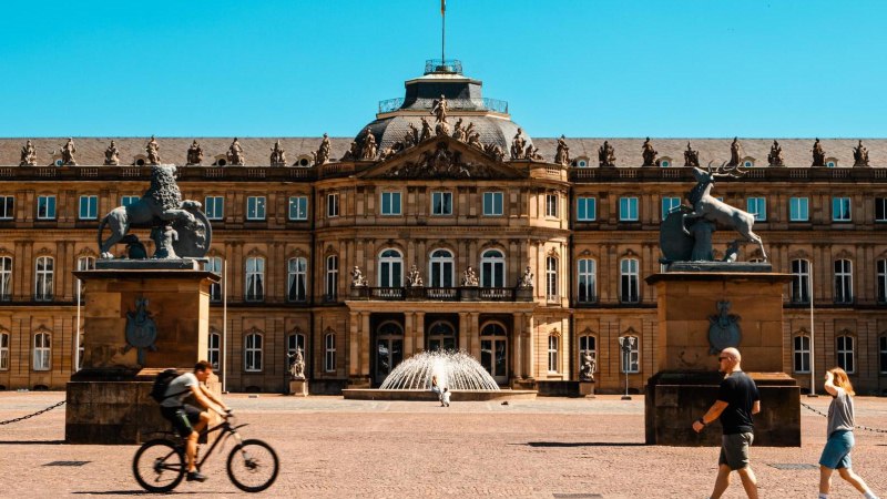 Das Neue Schloss Stuttgart bei sonnigem Wetter. Menschen gehen und ein Fahrradfahrer fährt im Vordergrund. Zwei Statuen flankieren den Eingang., © Stuttgart-Marketing GmbH, Sarah Schmid Das Neue Schloss Stuttgart bei sonnigem Wetter. Menschen gehen und ein Fahrradfahrer fährt im Vordergrund. Zwei Statuen flankieren den Eingang., © Stuttgart-Marketing GmbH, Sarah Schmid