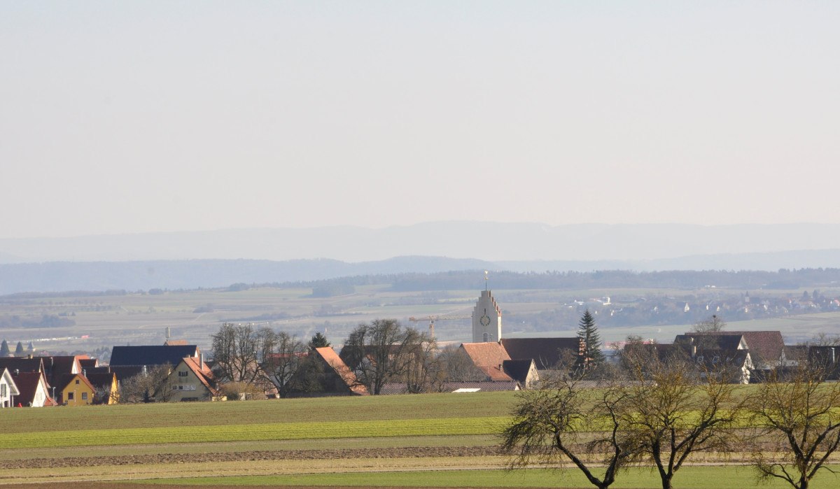 Blick auf ein Dorf mit Kirchturm in einer ländlichen Landschaft, umgeben von Feldern und Bäumen, im Hintergrund Hügel und blauer Himmel., © Natur.Nah. Schönbuch & Heckengäu Blick auf ein Dorf mit Kirchturm in einer ländlichen Landschaft, umgeben von Feldern und Bäumen, im Hintergrund Hügel und blauer Himmel., © Natur.Nah. Schönbuch & Heckengäu