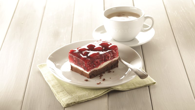 Himbeerschnitte auf einem Teller mit Löffel, daneben eine Tasse Kaffee auf einem Holztisch., © Bäckerei Veit, Victoria Huber Himbeerschnitte auf einem Teller mit Löffel, daneben eine Tasse Kaffee auf einem Holztisch., © Bäckerei Veit, Victoria Huber