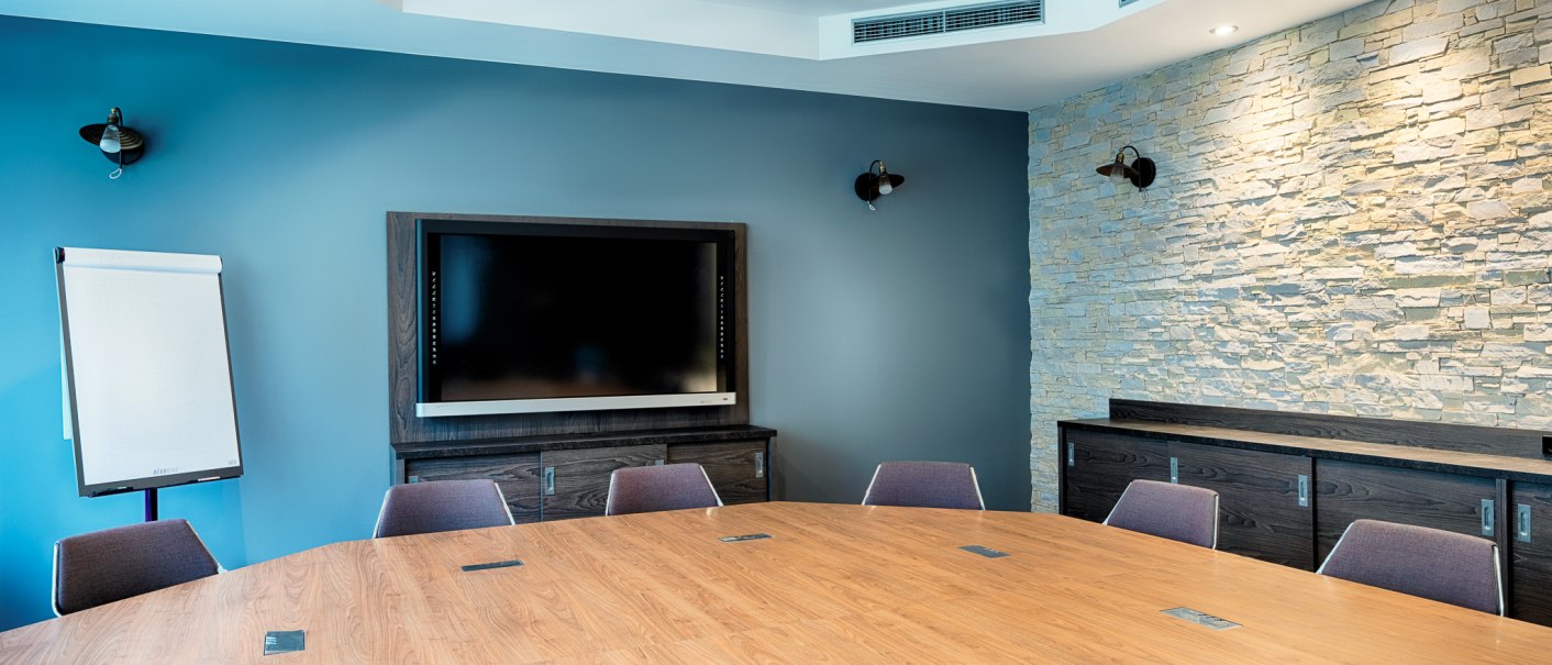 Moderner Tagungsraum mit großem Holztisch, Stühlen, einem Bildschirm an der Wand und einem Flipchart. Die Wände sind blau und mit Steinverkleidung., © Premier Inn Germany