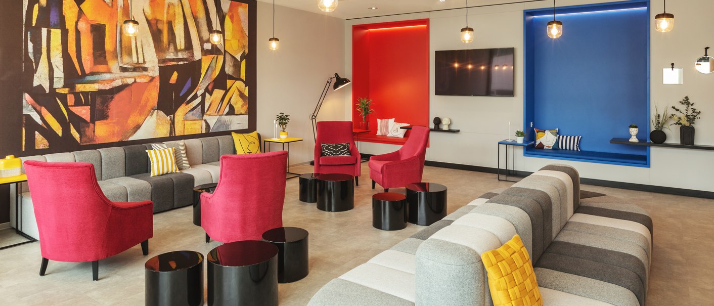 Moderne Lobby mit grauen und roten Sitzmöbeln, bunten Kissen, abstraktem Wandbild und farbigen Nischen in Rot und Blau., © the niu Form - Novum Hospitality