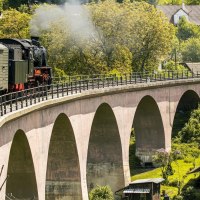 Eine Dampflokomotive zieht einen Zug &uuml;ber eine Br&uuml;cke in einer gr&uuml;nen, l&auml;ndlichen Landschaft. Menschen schauen aus den Fenstern und fotografieren., &copy; SMG, Sarah Schmid