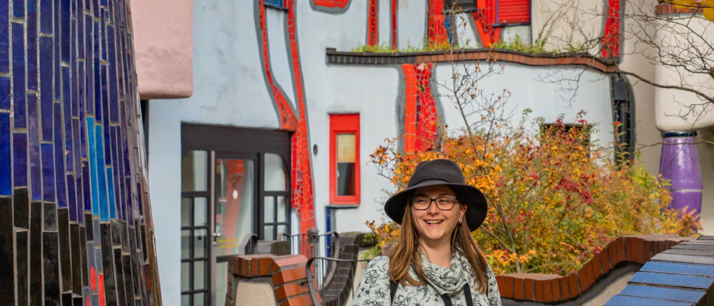 Frau mit Hut vor dem farbenfrohen Hundertwasserhaus in Plochingen, umgeben von herbstlichen Blättern., © Bildergalerie Attila