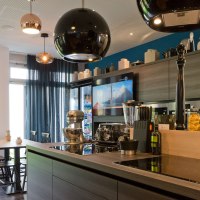 Moderne Küche mit Kochinsel, Esstisch, Hängelampen und Geräten. Große Fenster mit blauen Vorhängen lassen viel Licht herein., © Arthotel Ana / elaya