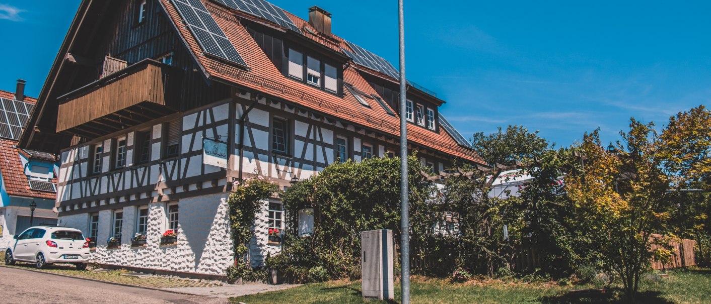 Fachwerkhaus mit Solarpanelen auf dem Dach, umgeben von Bäumen und einem Auto. Klare blaue Himmel und eine Straßenlaterne im Vordergrund., © Stadt Welzheim Fachwerkhaus mit Solarpanelen auf dem Dach, umgeben von Bäumen und einem Auto. Klare blaue Himmel und eine Straßenlaterne im Vordergrund., © Stadt Welzheim