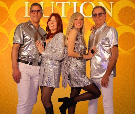 Vier Personen in glitzernden Outfits posieren vor einem gelben Hintergrund mit dem Schriftzug 'ABBAlution'., &copy; Touristik Bad Wildbad GmbH