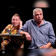 Zwei M&auml;nner sitzen an einem Tisch, einer tr&auml;gt ein buntes Hemd, der andere ein kariertes Hemd. Beide schauen in unterschiedliche Richtungen., &copy; links im Bild