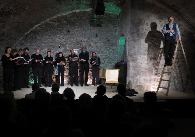 Ein Chor singt in einem Gewölbekeller. Ein Mann steht auf einer Leiter, während das Publikum zuschaut. Die Beleuchtung ist gedämpft., © Theater im Schlosskeller Ein Chor singt in einem Gewölbekeller. Ein Mann steht auf einer Leiter, während das Publikum zuschaut. Die Beleuchtung ist gedämpft., © Theater im Schlosskeller