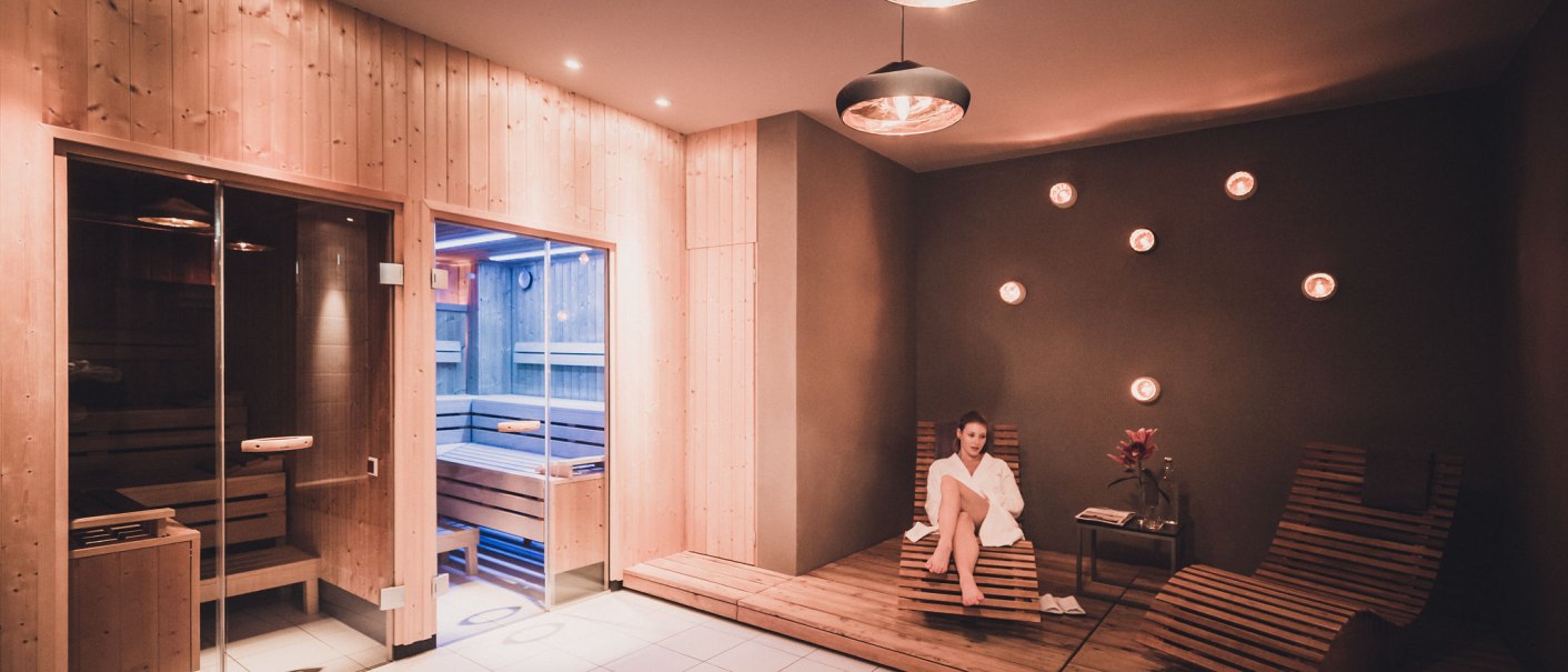 Moderner Saunabereich mit Holzverkleidung, zwei Saunakabinen, Liegestühlen und gedämpfter Beleuchtung. Eine Person sitzt entspannt auf einem Liegestuhl., © Abacco Hotels GmbH Moderner Saunabereich mit Holzverkleidung, zwei Saunakabinen, Liegestühlen und gedämpfter Beleuchtung. Eine Person sitzt entspannt auf einem Liegestuhl., © Abacco Hotels GmbH