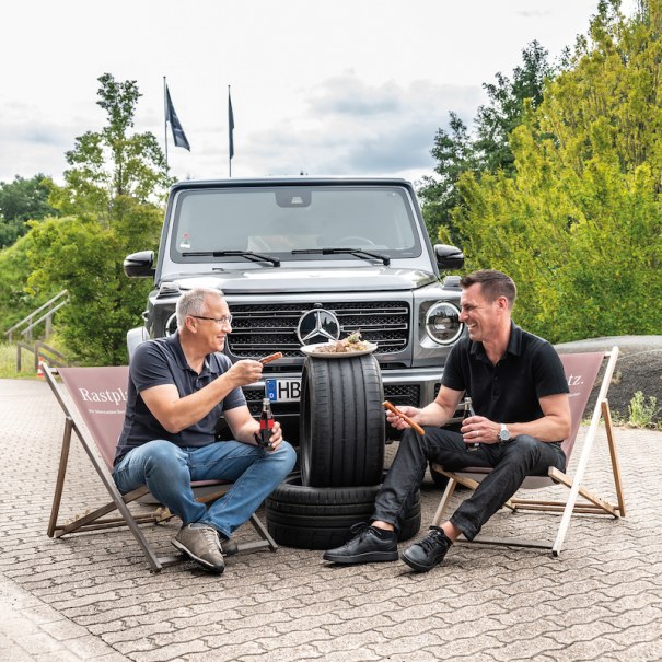 Zwei M&auml;nner sitzen auf Liegest&uuml;hlen vor einem Mercedes-Gel&auml;ndewagen und grillen auf einem Autoreifen. Sie lachen und genie&szlig;en die entspannte Atmosph&auml;re., &copy; Mercedes-Benz AG