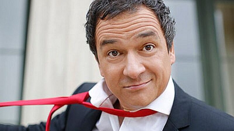 Ein Mann in einem Anzug zieht spielerisch an seiner roten Krawatte und schaut in die Kamera. Der Hintergrund ist unscharf., &copy; Stadthalle Leonberg