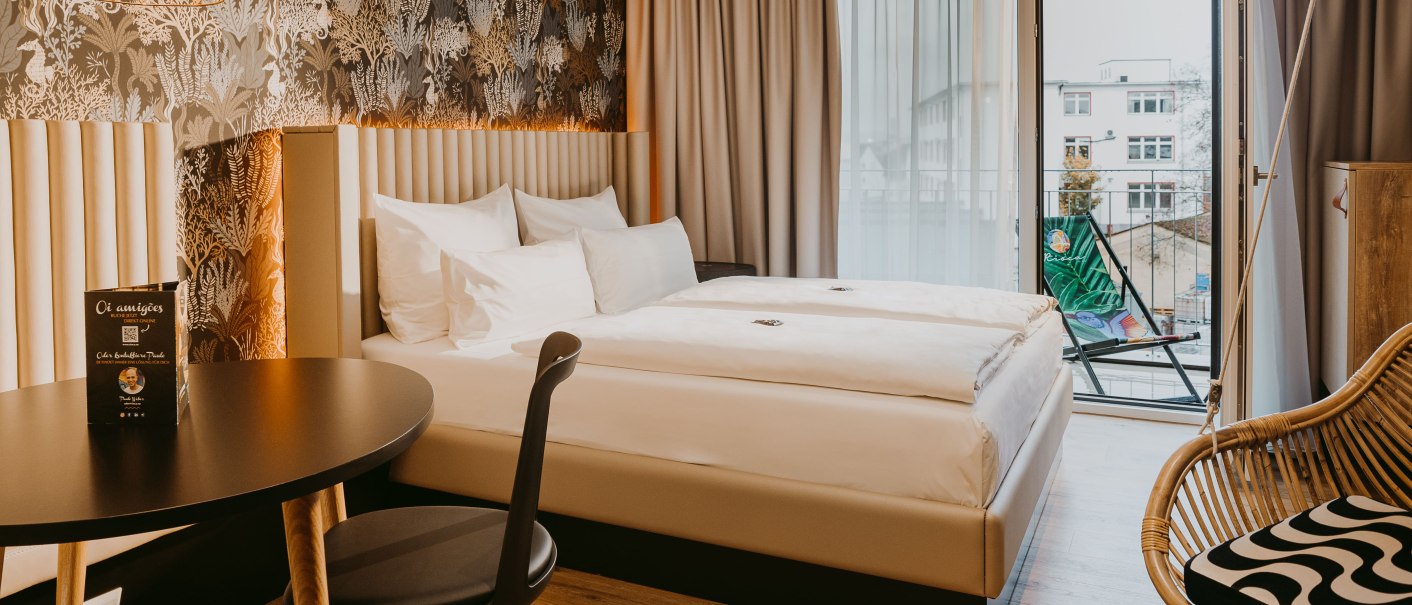 Modernes Hotelzimmer mit Doppelbett, stilvoller Tapete, Tisch und Stuhl. Zugang zu einem Balkon mit Liegestuhl. Helle, einladende Atmosphäre., © Rioca Stuttgart Posto 4 GmbH