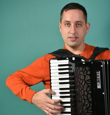 Ein Mann in einem orangefarbenen Pullover spielt Akkordeon vor einem türkisfarbenen Hintergrund. Er schaut in die Kamera., © Roman Dotsenko Ein Mann in einem orangefarbenen Pullover spielt Akkordeon vor einem türkisfarbenen Hintergrund. Er schaut in die Kamera., © Roman Dotsenko