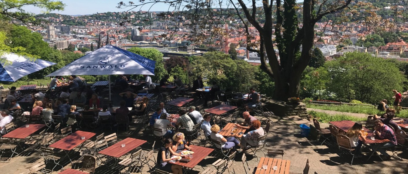 Ein Biergarten auf der Karlshöhe mit Blick auf Stuttgart. Menschen sitzen an Tischen unter Sonnenschirmen, umgeben von Bäumen und einer Stadtansicht., © Stuttgart-Marketing GmbH