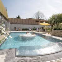 Außenbereich der Barbarossa-Thermen mit Pool, sprudelndem Wasser und Liegestühlen. Menschen entspannen unter Sonnenschirmen. Blumen und Bäume im Hintergrund., © Barbarossa-Thermen Göppingen