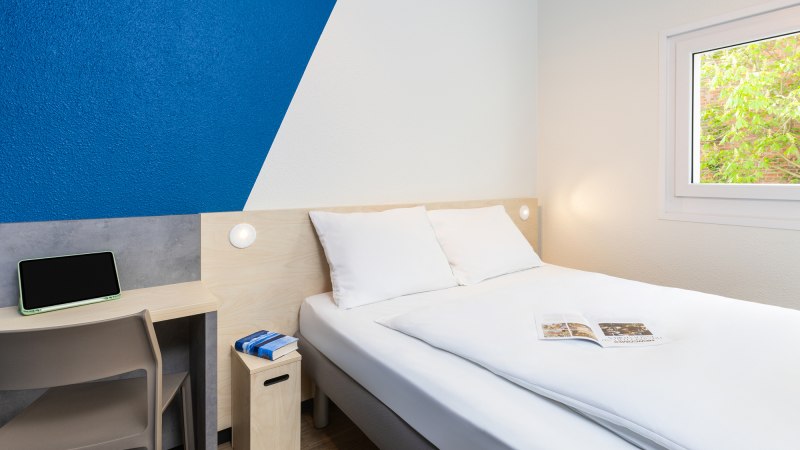 Ein schlichtes Hotelzimmer mit Doppelbett, blau-weißer Wand, kleinem Schreibtisch und Fenster mit Blick ins Grüne., © DQuadrat Living GmbH Ein schlichtes Hotelzimmer mit Doppelbett, blau-weißer Wand, kleinem Schreibtisch und Fenster mit Blick ins Grüne., © DQuadrat Living GmbH