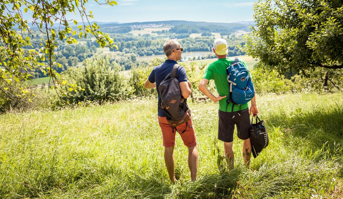Zwei Männer mit Rucksäcken stehen auf einer Wiese und schauen in die grüne Landschaft unter blauem Himmel., © hochgehberge