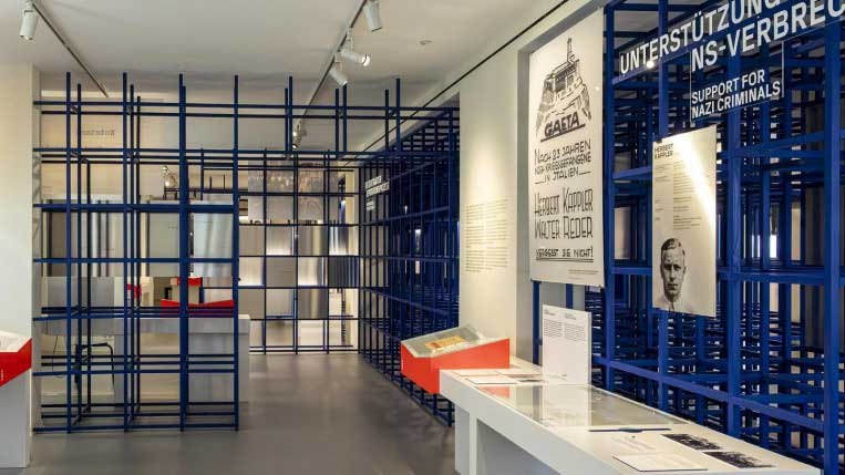 Ein Ausstellungsraum mit blauen Gitterstrukturen, historischen Plakaten und Informationsmaterialien an den Wänden., © Haus der Geschichte Baden-Württemberg