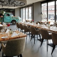 Ein modernes Restaurant mit Holztischen, eleganten Stühlen und einem alten blauen Truck als Dekoration. Große Fenster lassen viel Licht herein., © V8 Hotel, Frank Hoppe
