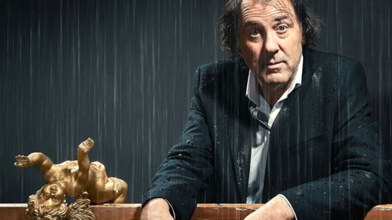 Ein Mann in einem nassen Anzug sitzt unter Regen. Neben ihm liegt eine goldene Engelsfigur auf einem Tisch., © Stadthalle Leonberg