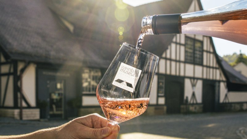 Ros&eacute;wein wird vor dem Weinbaumuseum in ein Glas eingeschenkt, im Hintergrund ein Fachwerkgeb&auml;ude im Sonnenlicht., &copy; SMG, Sarah Schmid