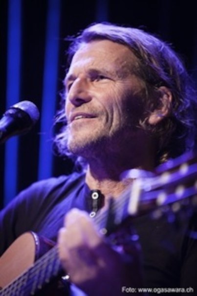 Ein Mann mit Gitarre singt auf einer Bühne, beleuchtet von blauem Licht. Er trägt ein schwarzes Shirt und hat längeres Haar., © SBEntertainment GmbH & Co. KG