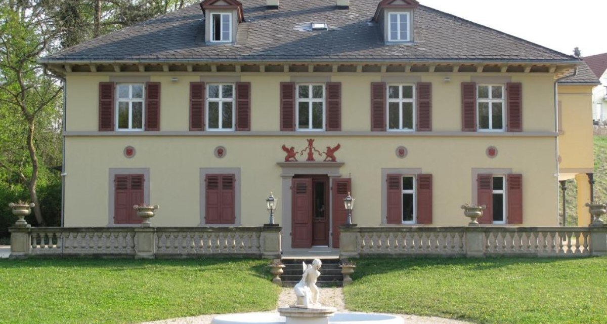 Gelbes zweistöckiges Haus mit roten Fensterläden, umgeben von Bäumen. Im Vorgarten steht eine Statue auf einem Brunnen., © Ludwigsburg - Stuttgart-Marketing GmbH Gelbes zweistöckiges Haus mit roten Fensterläden, umgeben von Bäumen. Im Vorgarten steht eine Statue auf einem Brunnen., © Ludwigsburg - Stuttgart-Marketing GmbH