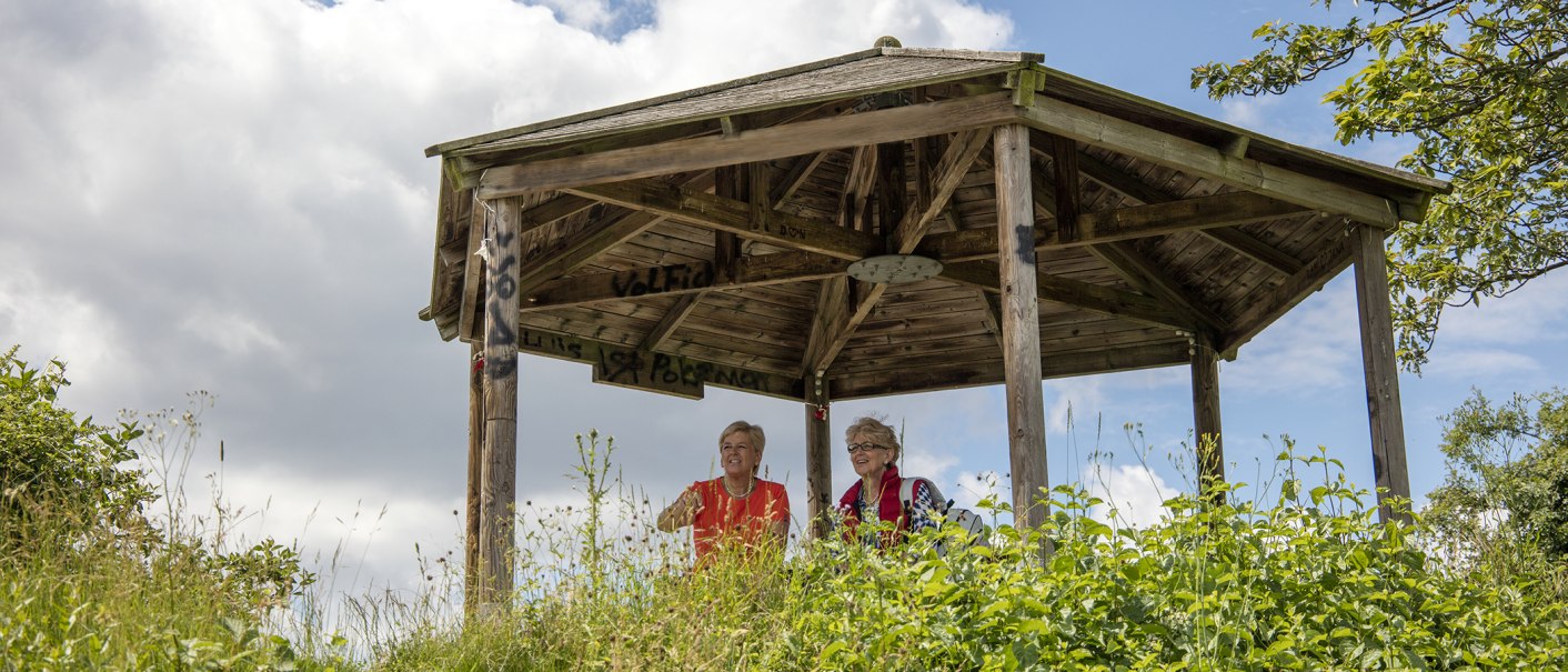 Zwei Personen sitzen in einem hölzernen Pavillon auf einem Hügel, umgeben von Pflanzen und bewölktem Himmel., © SMG, Achim Mende
