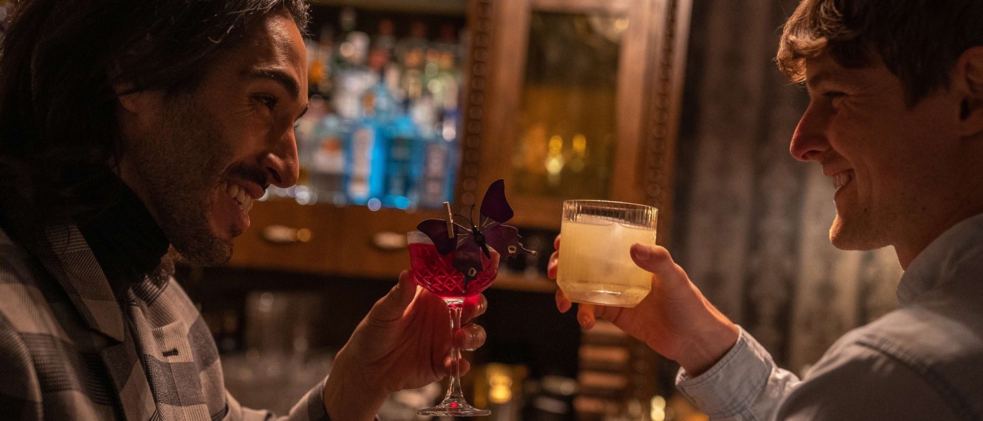 Zwei Männer stoßen in einer Bar mit Cocktails an, einer in einem karierten Sakko, der andere in einem hellen Hemd. Im Hintergrund sind Flaschen zu sehen., © SMG, Martina Denker