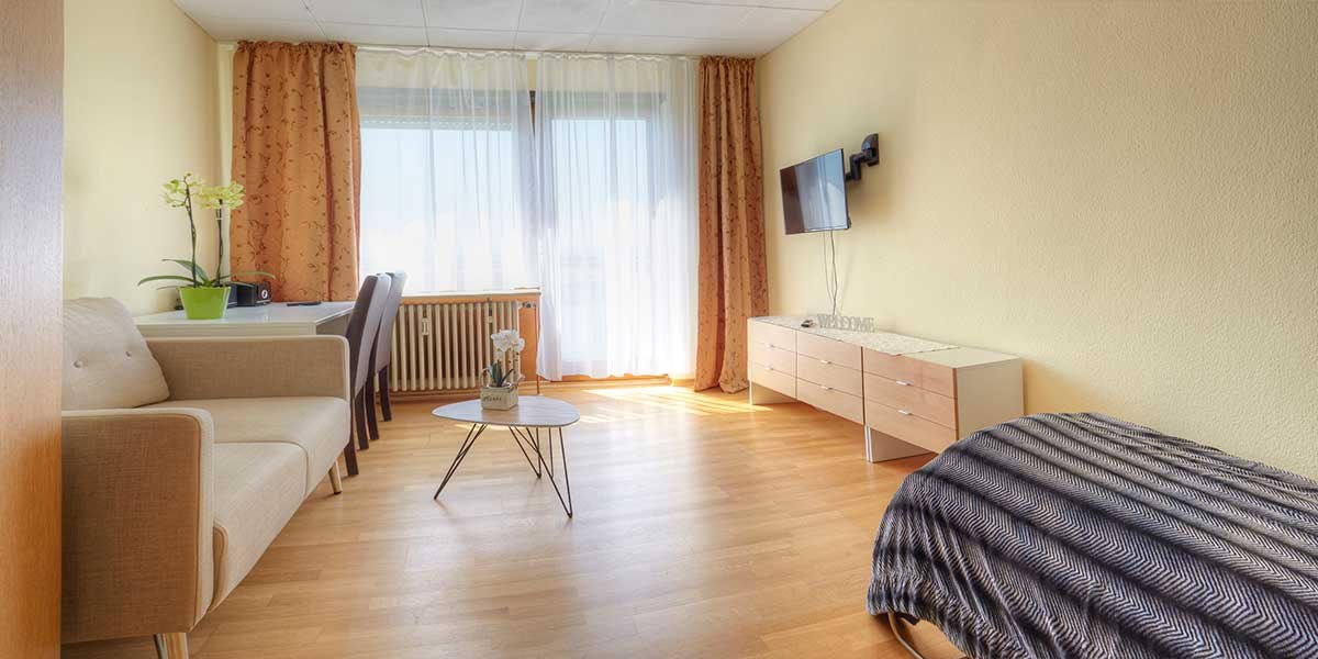 Helles Einzelzimmer-Apartment mit Sofa, Tisch, Schreibtisch, TV und Bett. Große Fenster mit Vorhängen lassen viel Licht herein., © Kemnater Hof GmbH