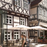 Belebte Straße mit Fachwerkhäusern, Cafés und Passanten. Menschen sitzen unter Sonnenschirmen, während andere vorbeigehen. Palmen schmücken die Szene., © Oswald Belebte Straße mit Fachwerkhäusern, Cafés und Passanten. Menschen sitzen unter Sonnenschirmen, während andere vorbeigehen. Palmen schmücken die Szene., © Oswald