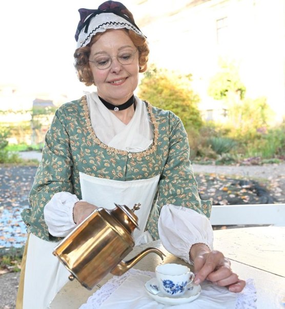 Eine Frau in historischer Kleidung gie&szlig;t Tee aus einer Kanne in eine Tasse. Sie tr&auml;gt eine Brille und l&auml;chelt. Im Hintergrund ist ein Garten zu sehen., &copy; Copyright: Tourismus & Events Ludwigsburg, Fotograf: Reiner Pfisterer