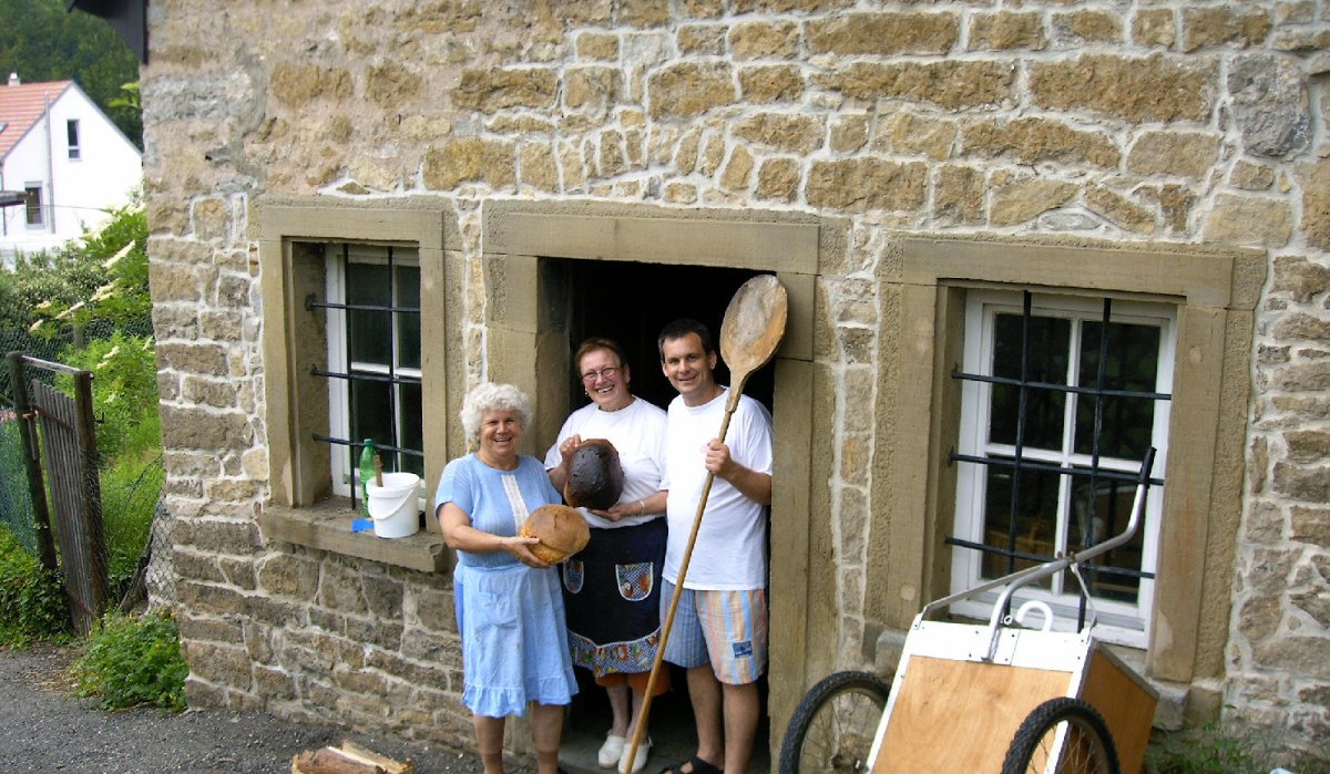 Drei Personen stehen vor einem alten Backhaus aus Stein, halten Brot und Backutensilien. Ein Handwagen lehnt an der Wand., © Land der 1000 Hügel - Kraichgau-Stromberg