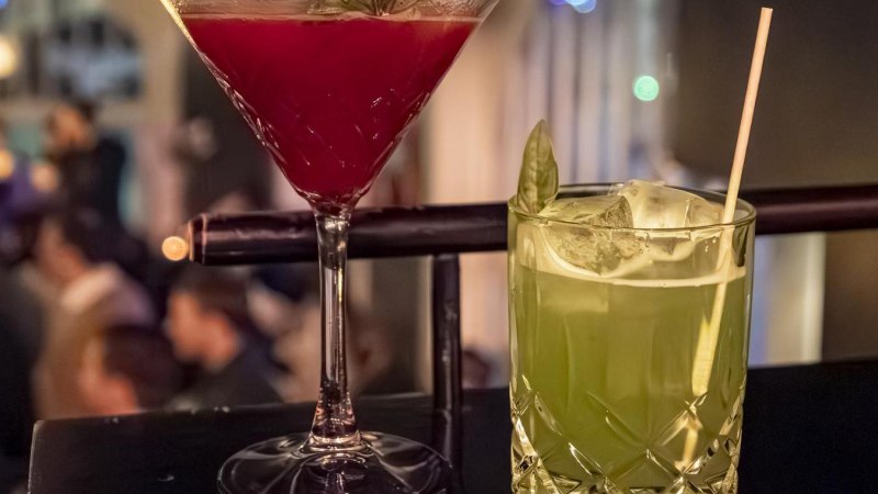 Zwei Cocktails auf einer Bar: ein roter im Martiniglas und ein gr&uuml;ner im Tumblerglas mit Strohhalm, im Hintergrund unscharfe Bar-Szene., &copy; SMG, Sarah Schmid
