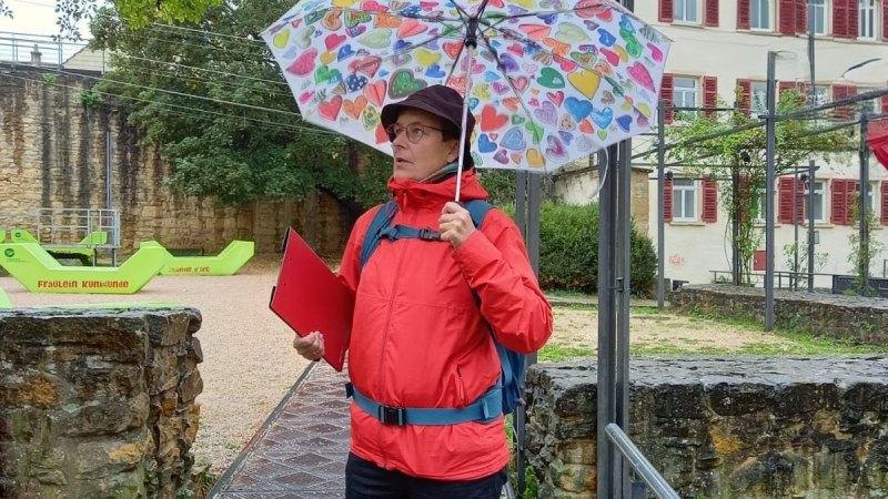 Eine Person mit roter Jacke und buntem Regenschirm steht auf einem Gehweg. Im Hintergrund sind B&auml;ume und ein Geb&auml;ude mit roten Fensterl&auml;den zu sehen., &copy; Christa Schulthei&szlig;
