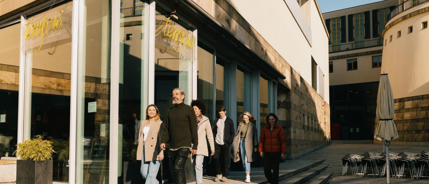 Eine Gruppe von Menschen verlässt das Tempus Café & Bistro, ein modernes Gebäude mit Glasfassade. Im Vordergrund stehen gestapelte Stühle und ein geschlossener Sonnenschirm., © Stuttgart-Marketing GmbH, Alwin Maigler