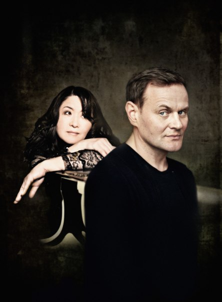 Eine Frau lehnt sich auf einen Tisch, während ein Mann im Vordergrund steht. Beide blicken in unterschiedliche Richtungen, vor einem dunklen Hintergrund., © Arens Schult