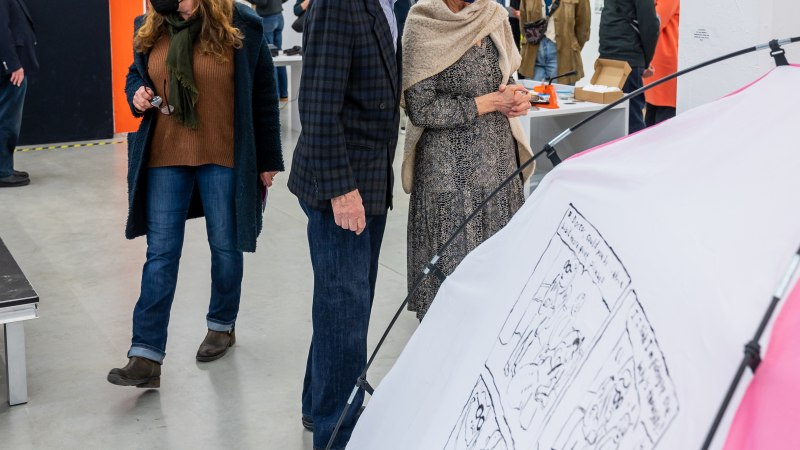 Besucher in einer Kunstgalerie betrachten eine Ausstellung. Sie tragen Masken und stehen vor einem Zelt mit Zeichnungen. Die Galerie hat bunte Säulen., © Kunsthalle Göppingen