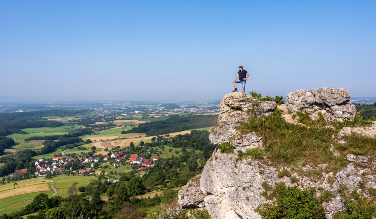 Eine Person steht auf einem Felsen und blickt auf eine weite, grüne Landschaft mit Feldern und einem Dorf im Hintergrund., © TMBW