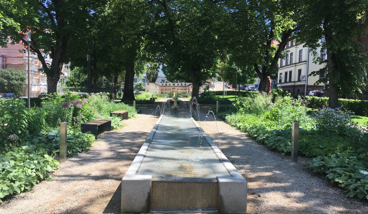Ein schattiger Park mit einem langen Wasserbecken, umgeben von Bäumen, Bänken und Blumenbeeten., © Touristik und Marketing GmbH Ein schattiger Park mit einem langen Wasserbecken, umgeben von Bäumen, Bänken und Blumenbeeten., © Touristik und Marketing GmbH