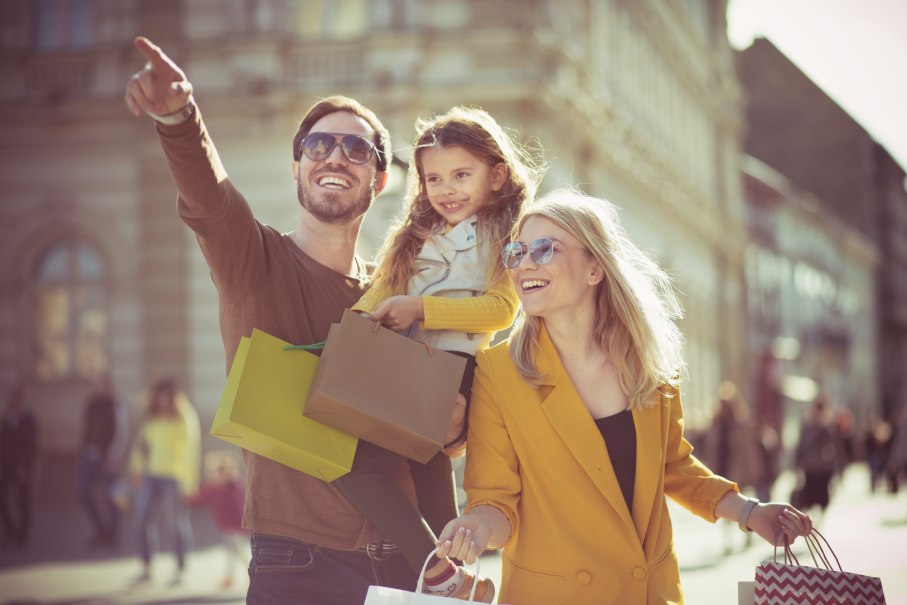 Eine fröhliche Familie mit Einkaufstüten genießt einen sonnigen Tag in der Stadt. Der Mann zeigt auf etwas in der Ferne.