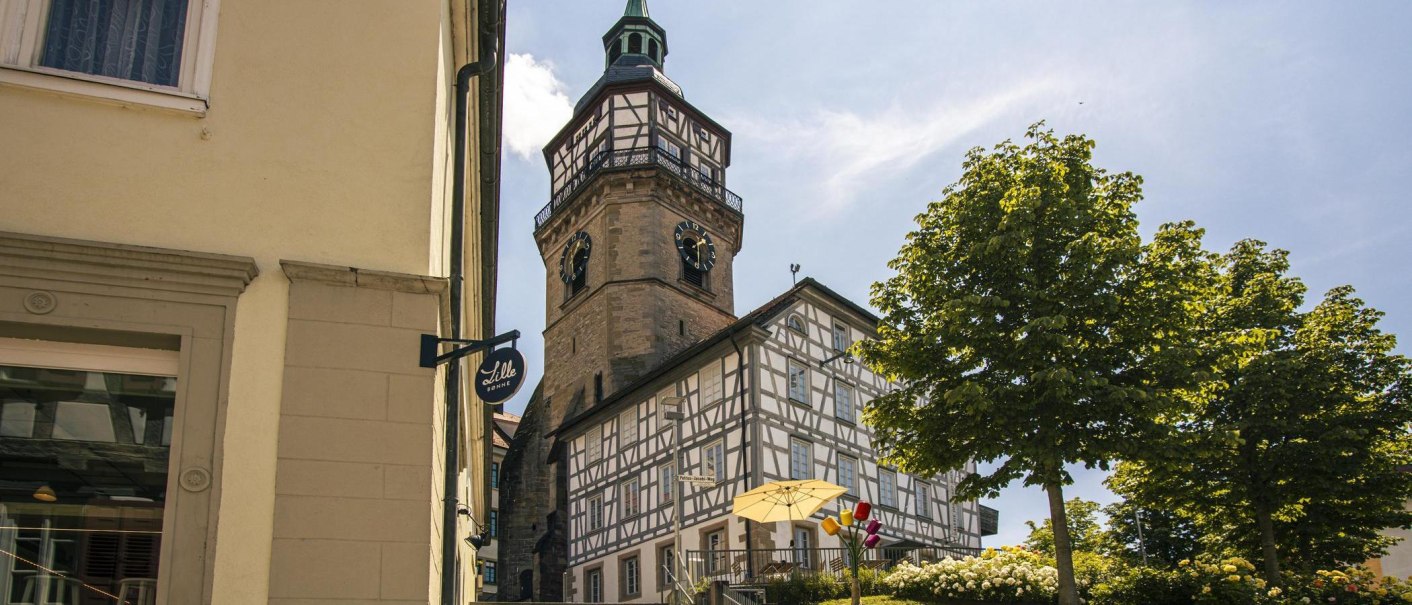 Fachwerkhaus und Kirchturm in Backnang, umgeben von Bäumen und Blumen, bei sonnigem Wetter. Ein Schild mit der Aufschrift 'Lille Bonne' ist sichtbar., © Stuttgart-Marketing GmbH, Sarah Schmid Fachwerkhaus und Kirchturm in Backnang, umgeben von Bäumen und Blumen, bei sonnigem Wetter. Ein Schild mit der Aufschrift 'Lille Bonne' ist sichtbar., © Stuttgart-Marketing GmbH, Sarah Schmid